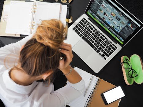 Overhead view of a stressed woman working at a desk with a weak laptop, phone, and notebooks.