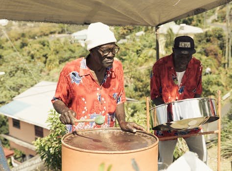 Two musicians play steel drums in a tropical outdoor setting with vibrant shirts and hats.