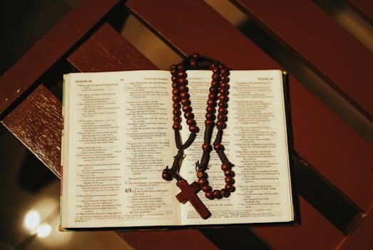 An open Bible with a wooden rosary lays on a wooden bench in a serene setting.