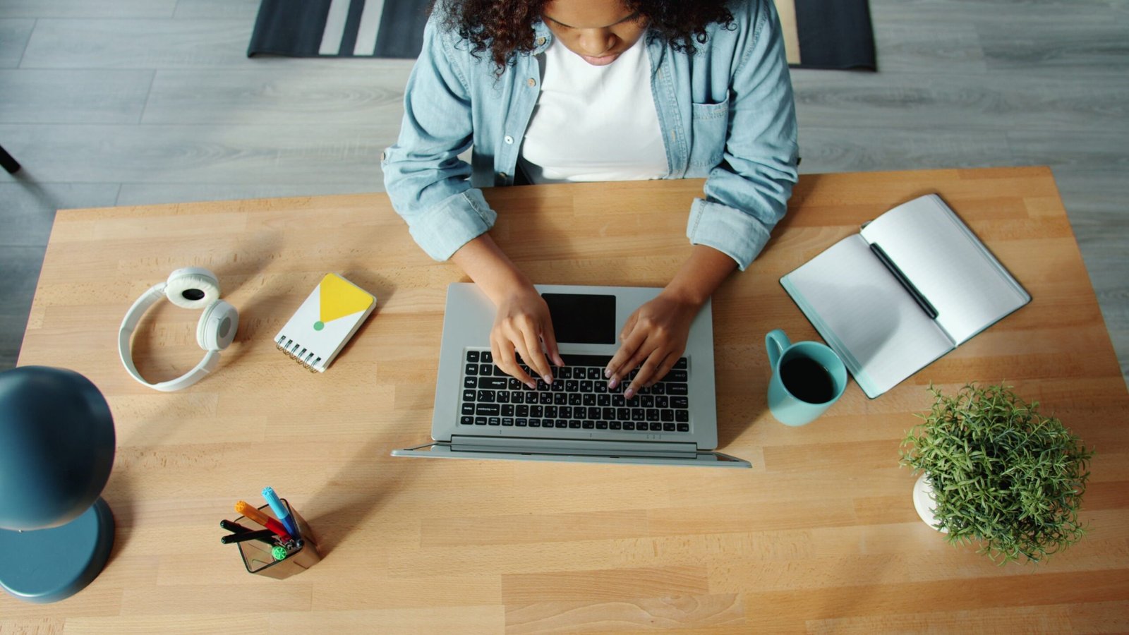 A woman using a laptop at a wooden desk with headphones, a notebook, and a plant.